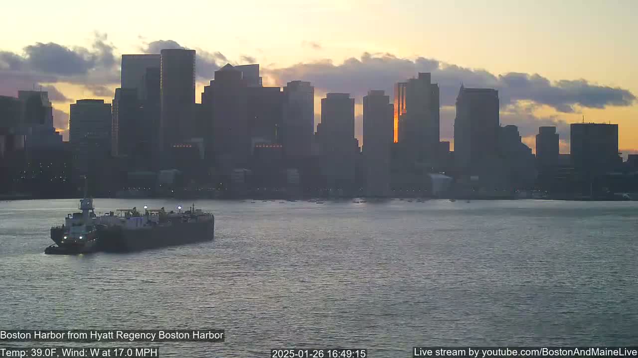 A tugboat pushes a barge across a calm harbor at sunset, with a city skyline silhouetted against a cloudy sky.