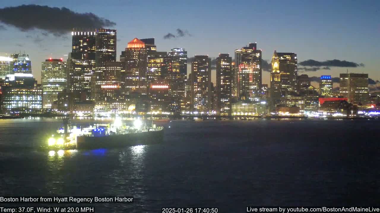 A brightly lit city skyline is seen across a dark body of water at night, under partly cloudy skies, with a tugboat visible in the foreground.