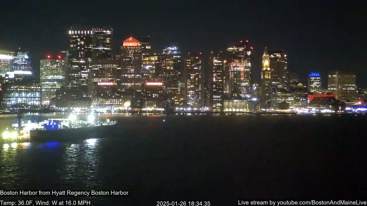 A brightly lit city skyline is seen across a dark body of water at night, under a clear sky with a 16 mph west wind and a temperature of 36°F.
