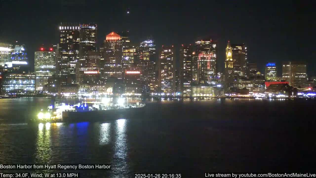A nighttime view of a brightly lit city skyline across a calm body of water, with a vessel visible in the foreground.