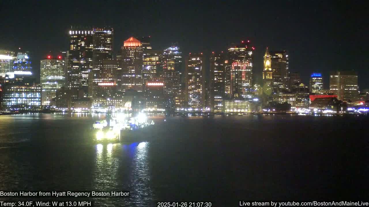 A nighttime view of a brightly lit city skyline across a dark body of water, with a vessel visible in the foreground under a clear sky and a reported wind of 13 mph.