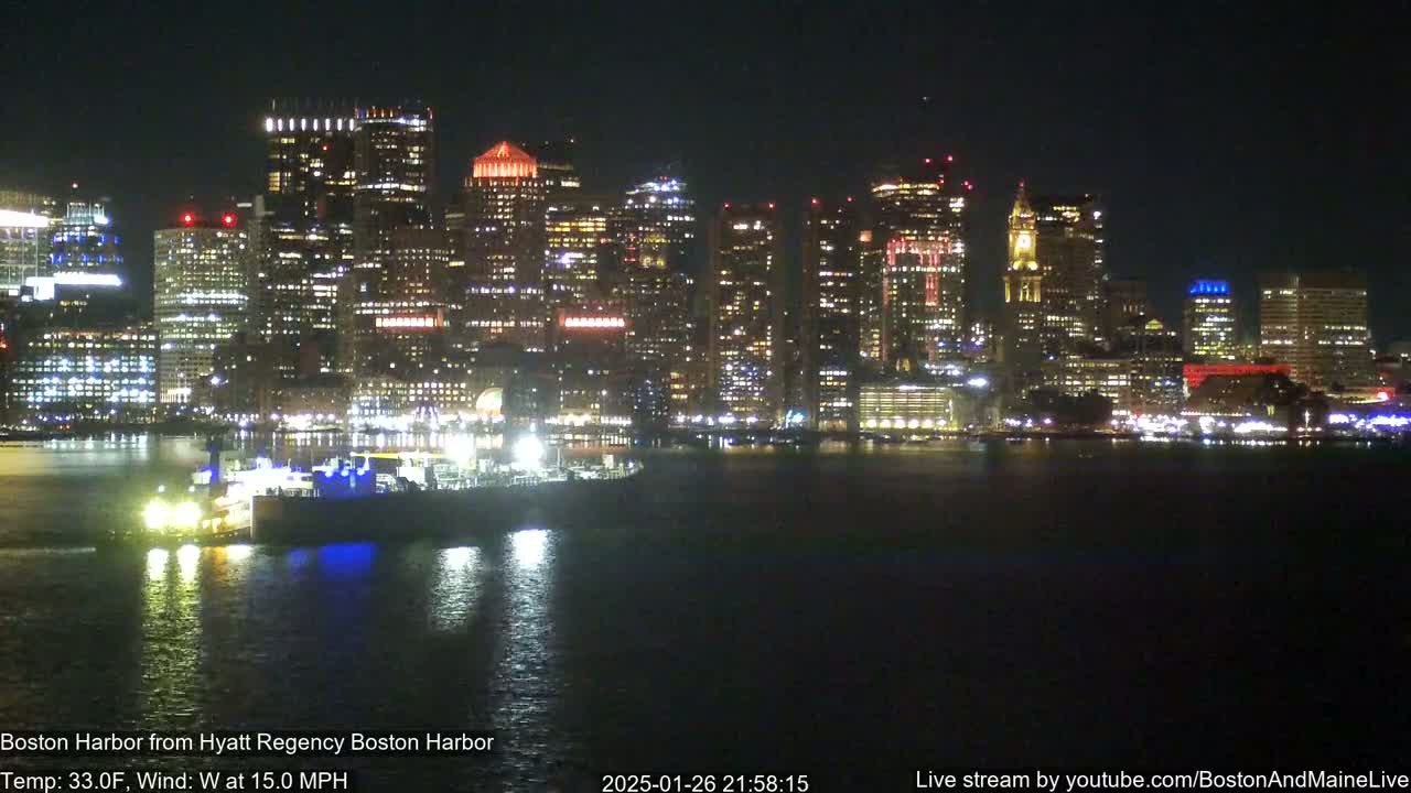 A nighttime view of a brightly lit city skyline across a dark body of water, with a vessel visible in the foreground under a clear sky.