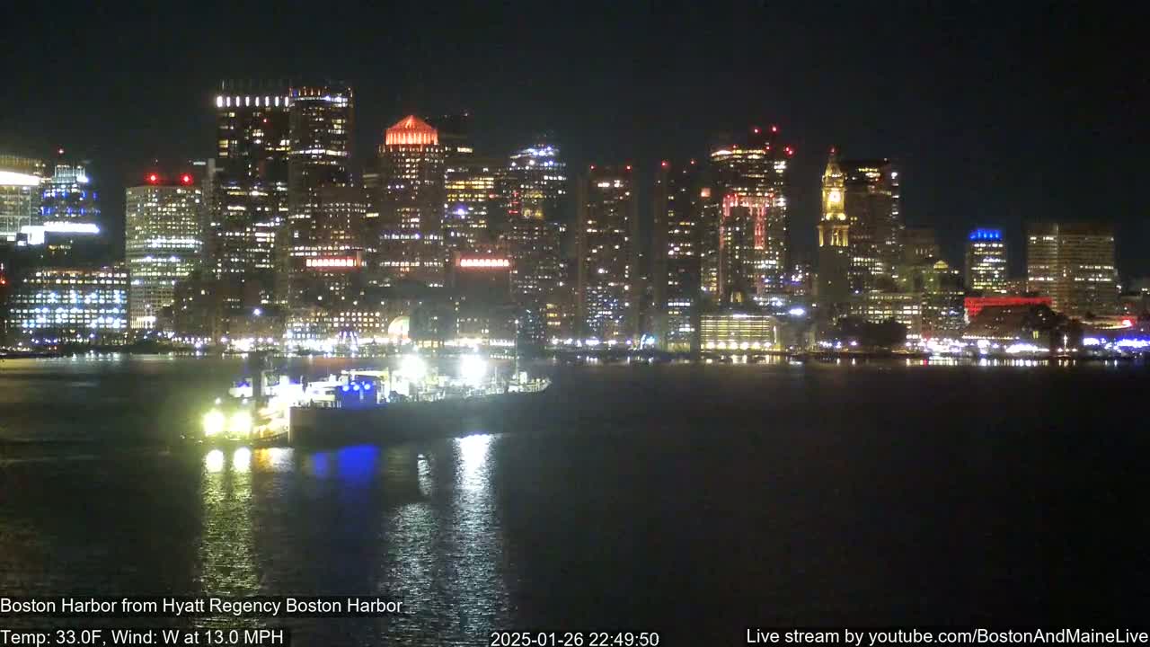 A nighttime view of a brightly lit city skyline across a dark body of water, with a vessel visible in the foreground under a calm, clear sky.