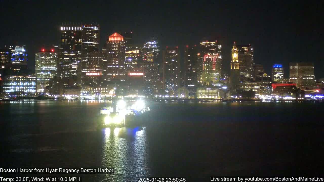 A brightly lit city skyline is seen across a dark body of water at night, with a vessel in the foreground and a 10 mph west wind.