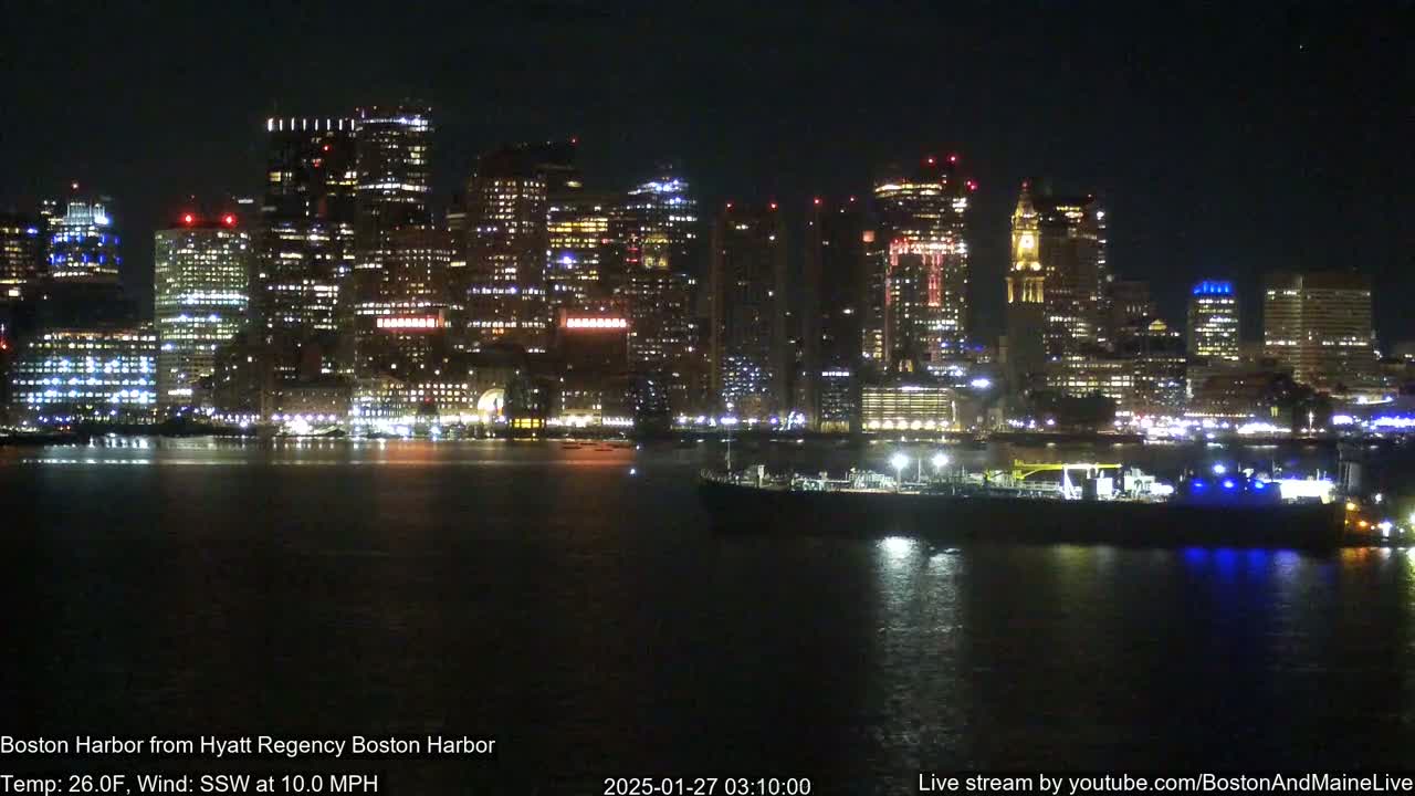 A nighttime view of a brightly lit city skyline across a dark harbor, with a large vessel visible in the water, under a clear sky with a 10 mph southwest wind and a temperature of 26 degrees Fahrenheit.
