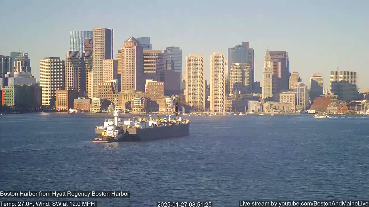 A barge is towed across a calm harbor in front of a city skyline under a clear, sunny sky.
