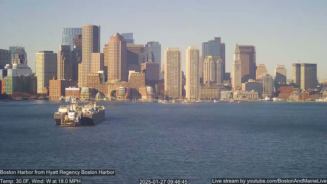 A barge is seen in the harbor in front of the Boston skyline on a sunny day with a west wind at 18 mph and a temperature of 30°F.