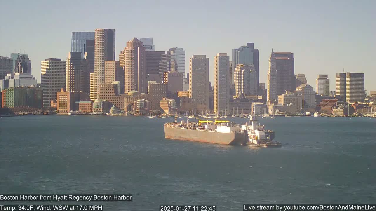 A barge is being towed across a harbor in front of a city skyline under a clear, sunny sky with a WSW wind at 17.0 mph.