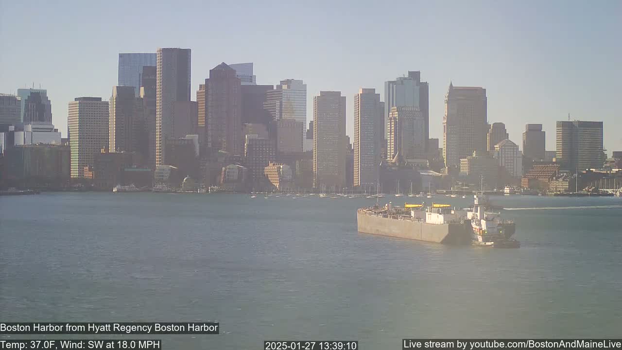 A barge is traveling on a harbor with a city skyline in the background under a clear, sunny sky.