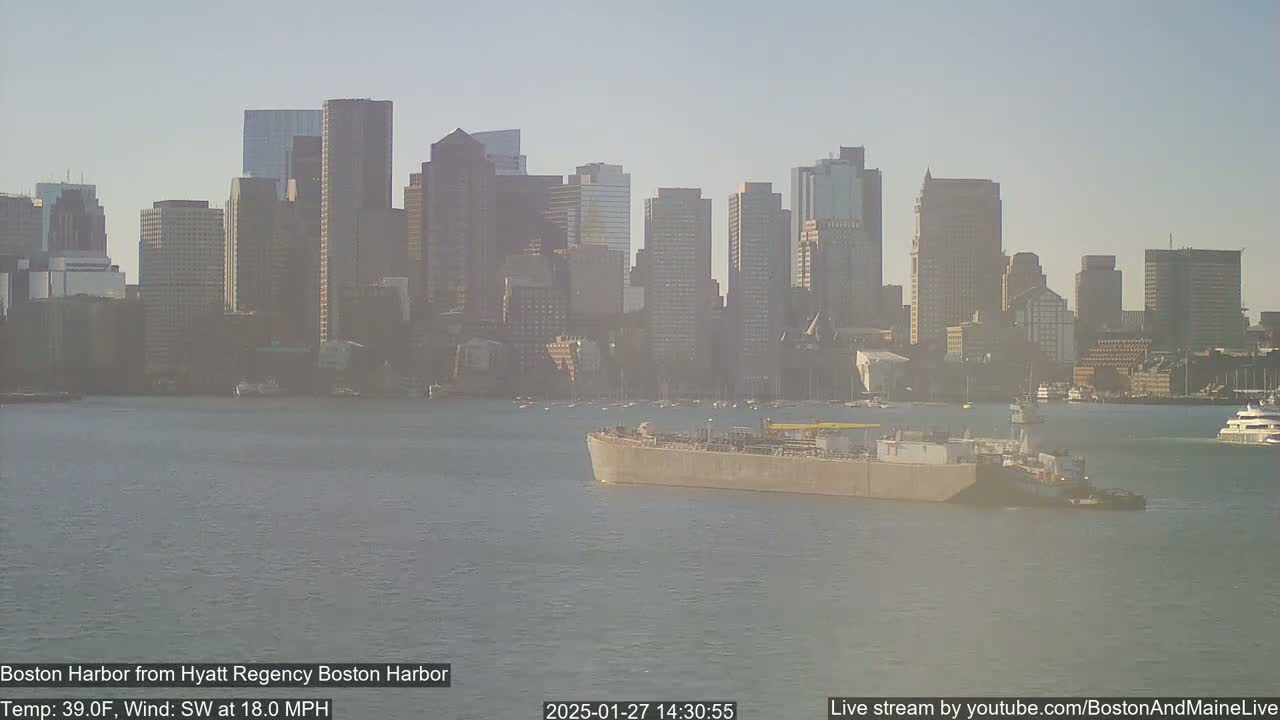 A barge is traveling on a calm harbor with a city skyline in the background under hazy conditions.