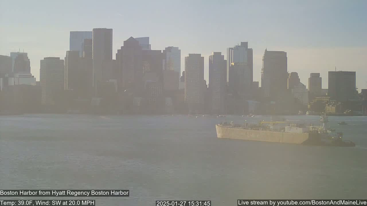 A barge is being towed across a harbor with a city skyline visible in the background under hazy, sunny conditions and a 20 mph southwest wind.