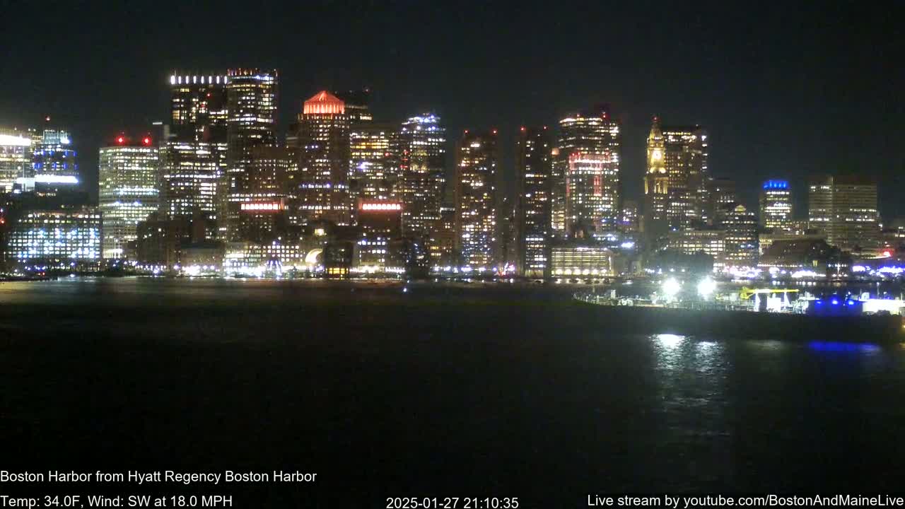 A brightly lit city skyline at night is seen across a dark body of water under a clear sky, with a southwest wind blowing at 18 mph and a temperature of 34°F.