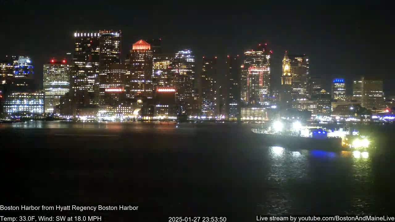 A brightly lit city skyline is seen across a dark body of water at night, with a large vessel in the foreground and a southwest wind at 18 mph.