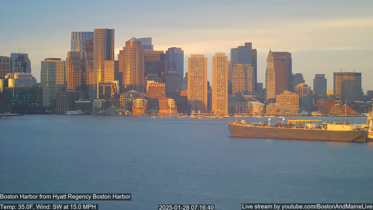 A large cargo ship sits in a calm harbor at sunset, with a city skyline in the background under a clear, pale sky.