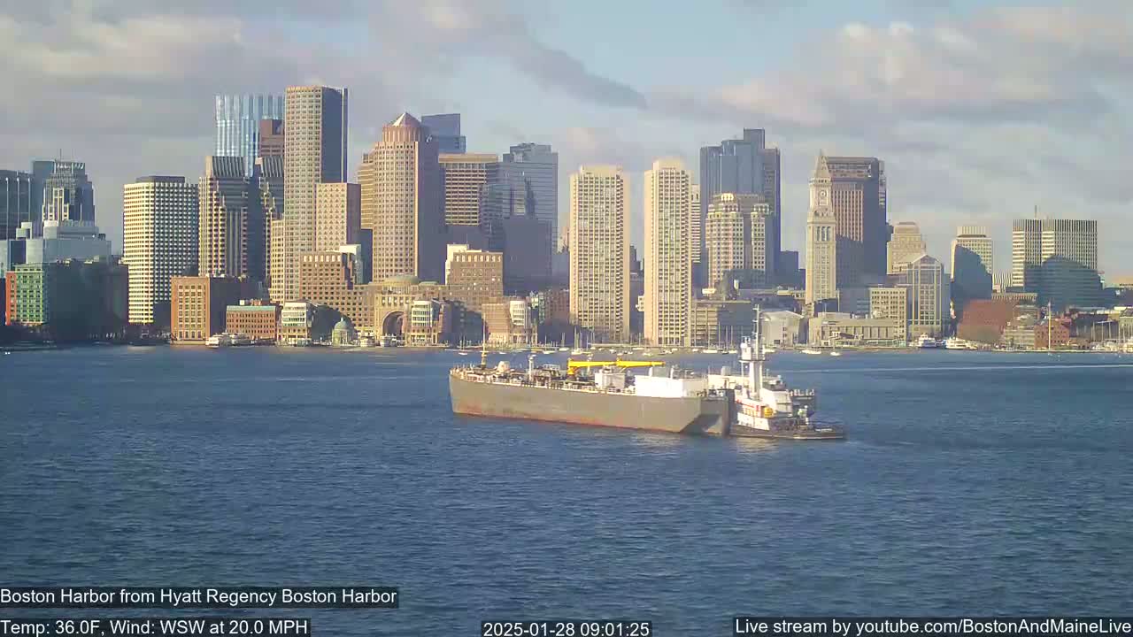 A large barge is being towed across a harbor in front of a city skyline under partly cloudy skies with a WSW wind at 20 mph.