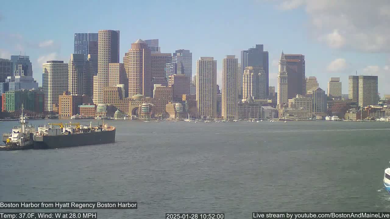 A large barge is towed across a choppy harbor in front of a city skyline on a partly cloudy day with a 28 mph west wind.