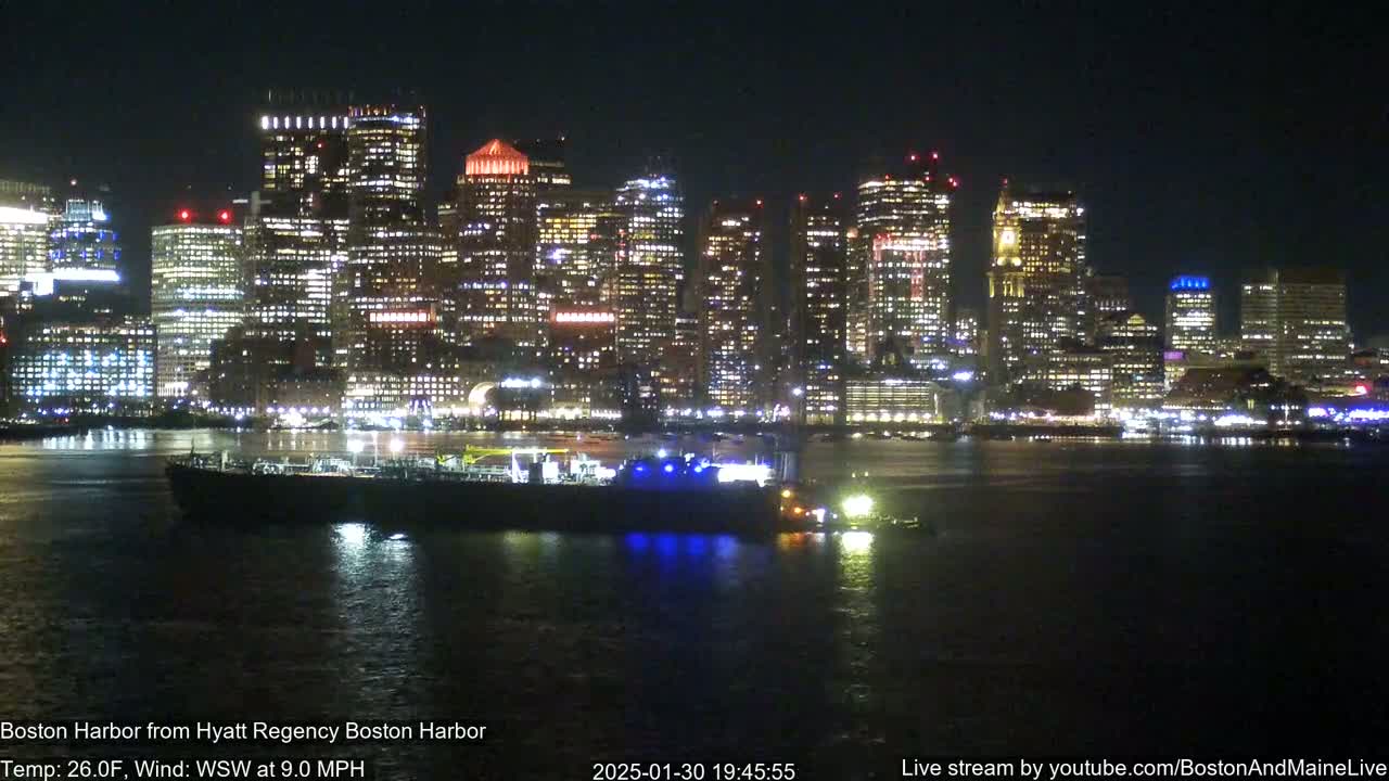 A brightly lit city skyline at night is seen across a dark body of water, with a large vessel in the foreground.
