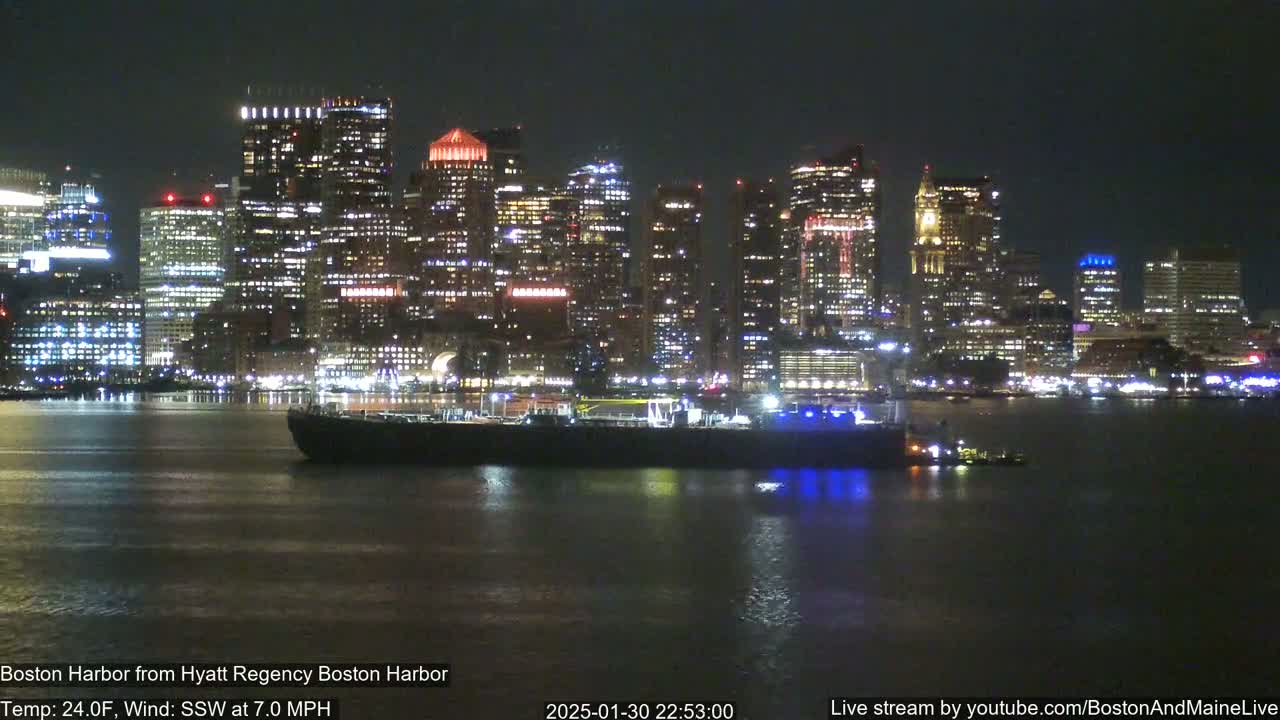 A brightly lit city skyline is reflected in dark water at night, with a barge and tugboat visible in the foreground, under a clear, cool sky.
