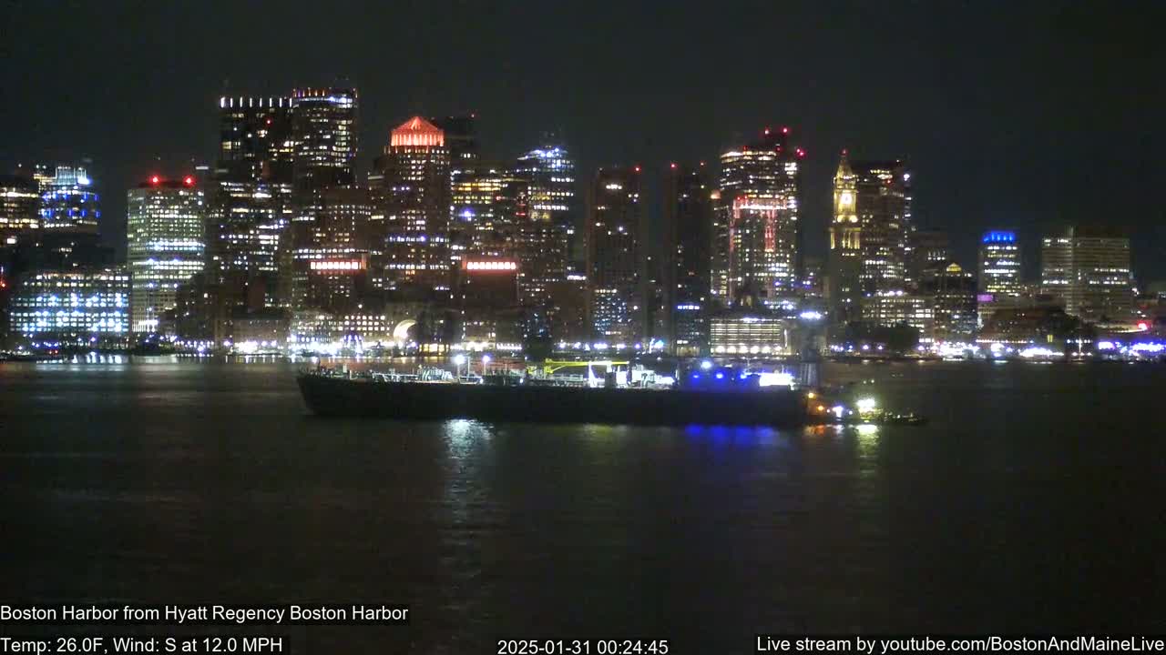A brightly lit city skyline is seen across a dark body of water at night, with a large vessel and smaller tugboat in the foreground, under a clear sky with a south wind.