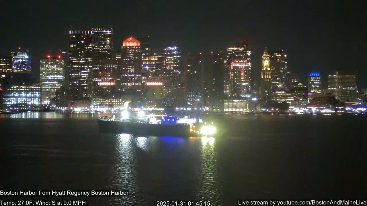 A nighttime view of a brightly lit city skyline across a dark body of water, with a large vessel near the foreground, under a clear sky.
