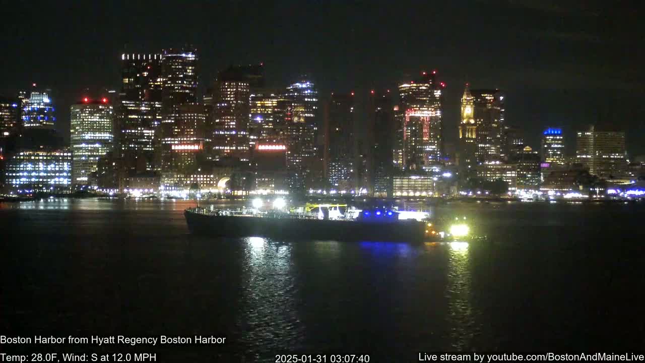A nighttime view of a brightly lit city skyline across a dark body of water with a ship in the foreground, under a clear sky with a south wind at 12 mph.