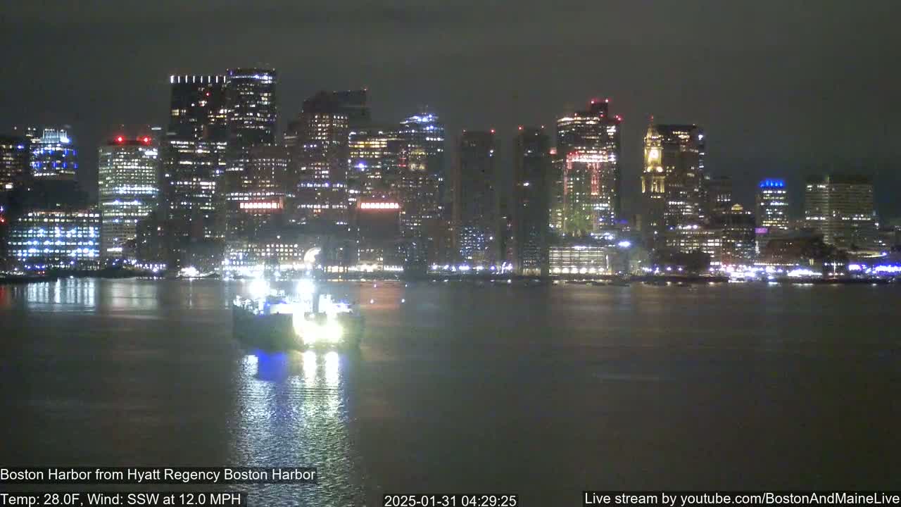 A nighttime view of a brightly lit city skyline across a dark body of water, with a vessel in the foreground and a south-southwesterly wind at 12 mph.
