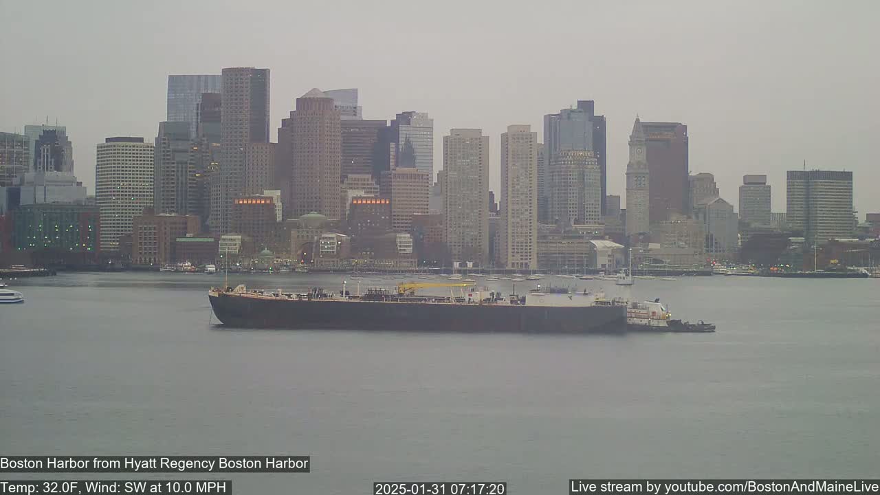 A large barge is being towed on calm, gray water in front of a city skyline under an overcast sky.