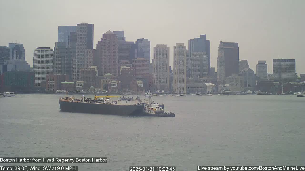 A barge being towed by a tugboat is seen in a harbor with a hazy cityscape in the background, under overcast conditions with a southwest wind.
