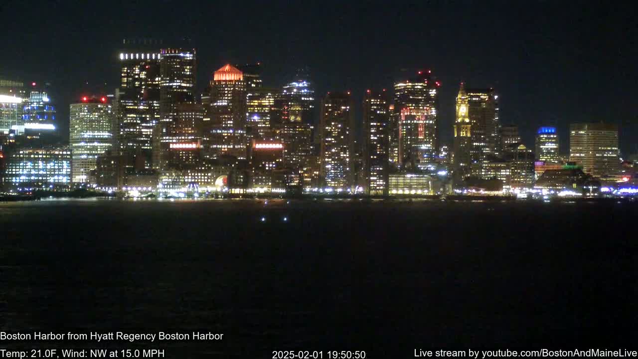 A brightly lit cityscape is seen across a dark body of water at night, under a clear sky with a northwest wind at 15 mph and a temperature of 21°F.