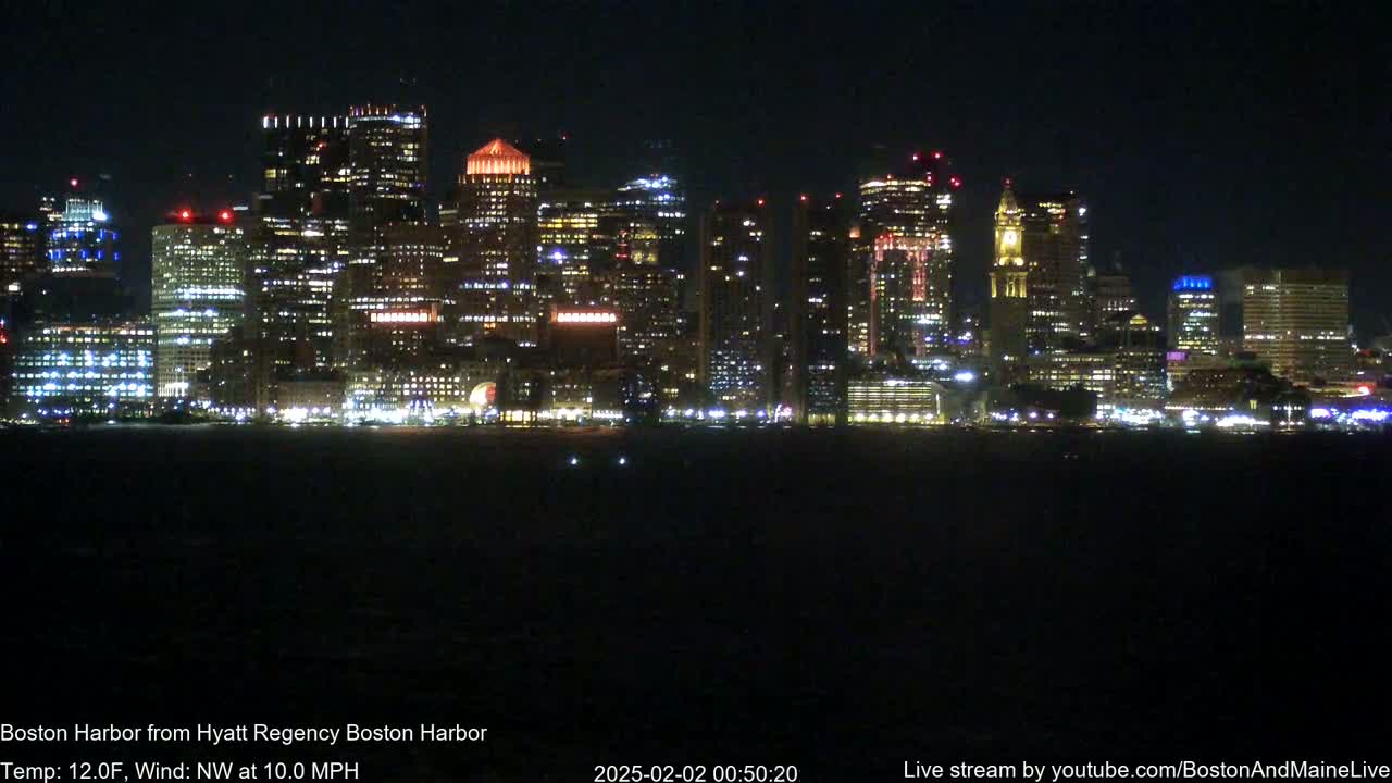 A brightly lit city skyline is seen across a dark body of water at night, with a northwest wind blowing at 10 mph and a temperature of 12°F.