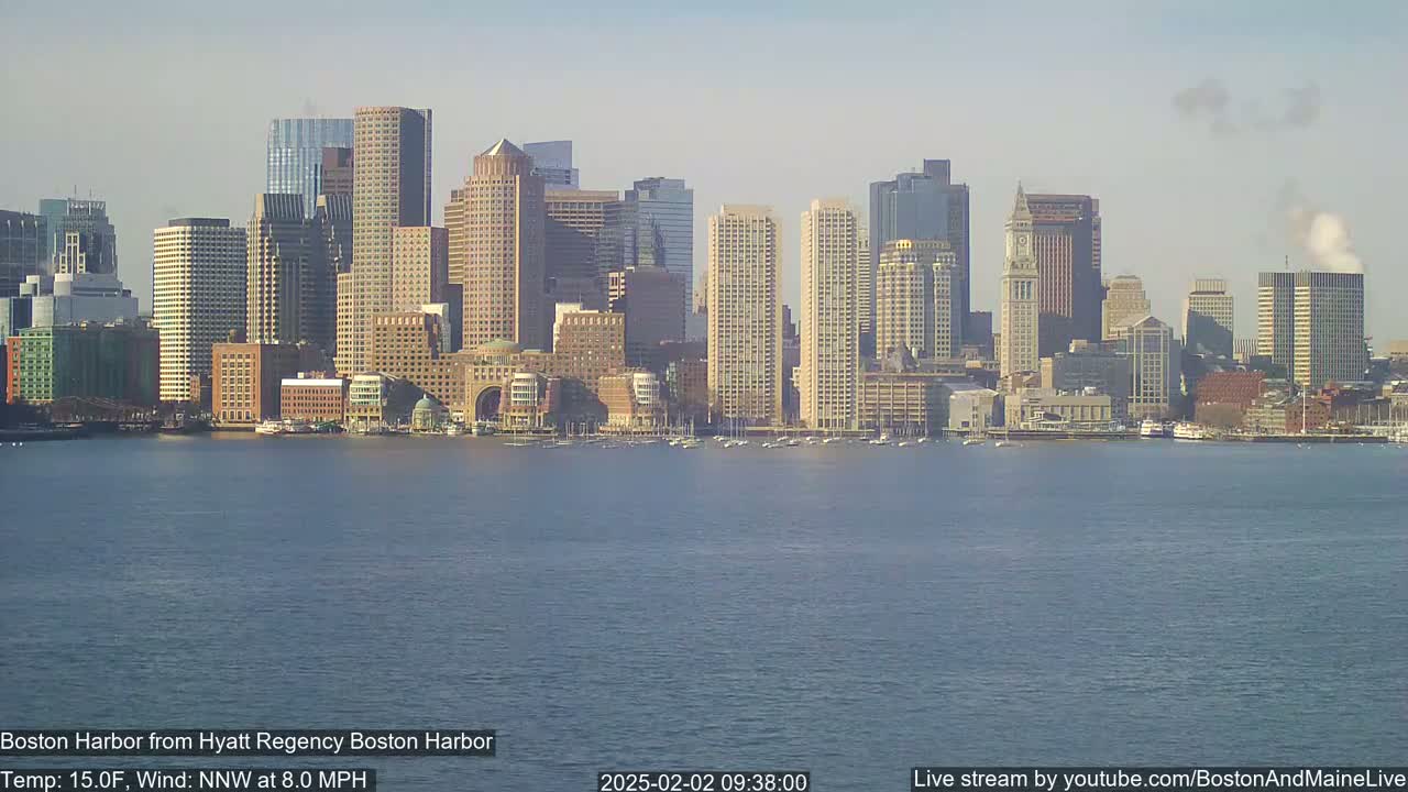 A cityscape of numerous tall buildings sits along a calm body of water under a hazy sky.