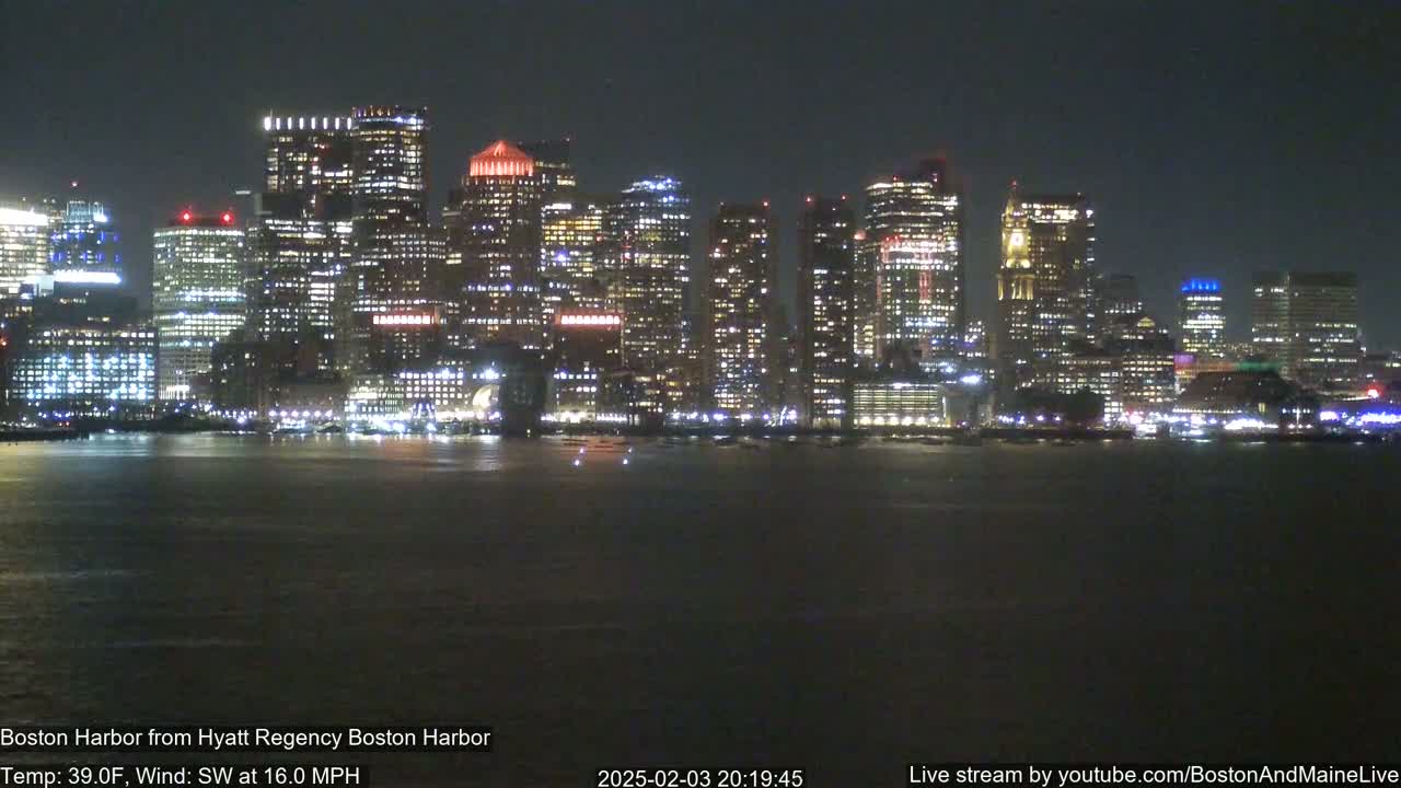 A brightly lit cityscape is seen across a dark body of water at night, under a clear sky with a southwest wind at 16 mph.