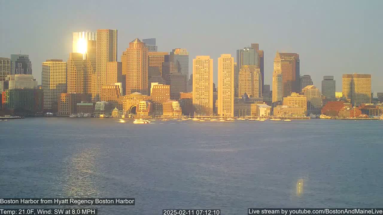 A city skyline, illuminated by the setting sun, is seen across a calm harbor.