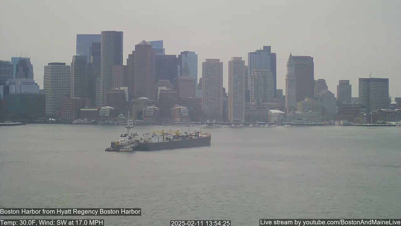A barge is on the water in front of a hazy city skyline under a gray sky with a southwest wind at 17 mph and a temperature of 30 degrees Fahrenheit.