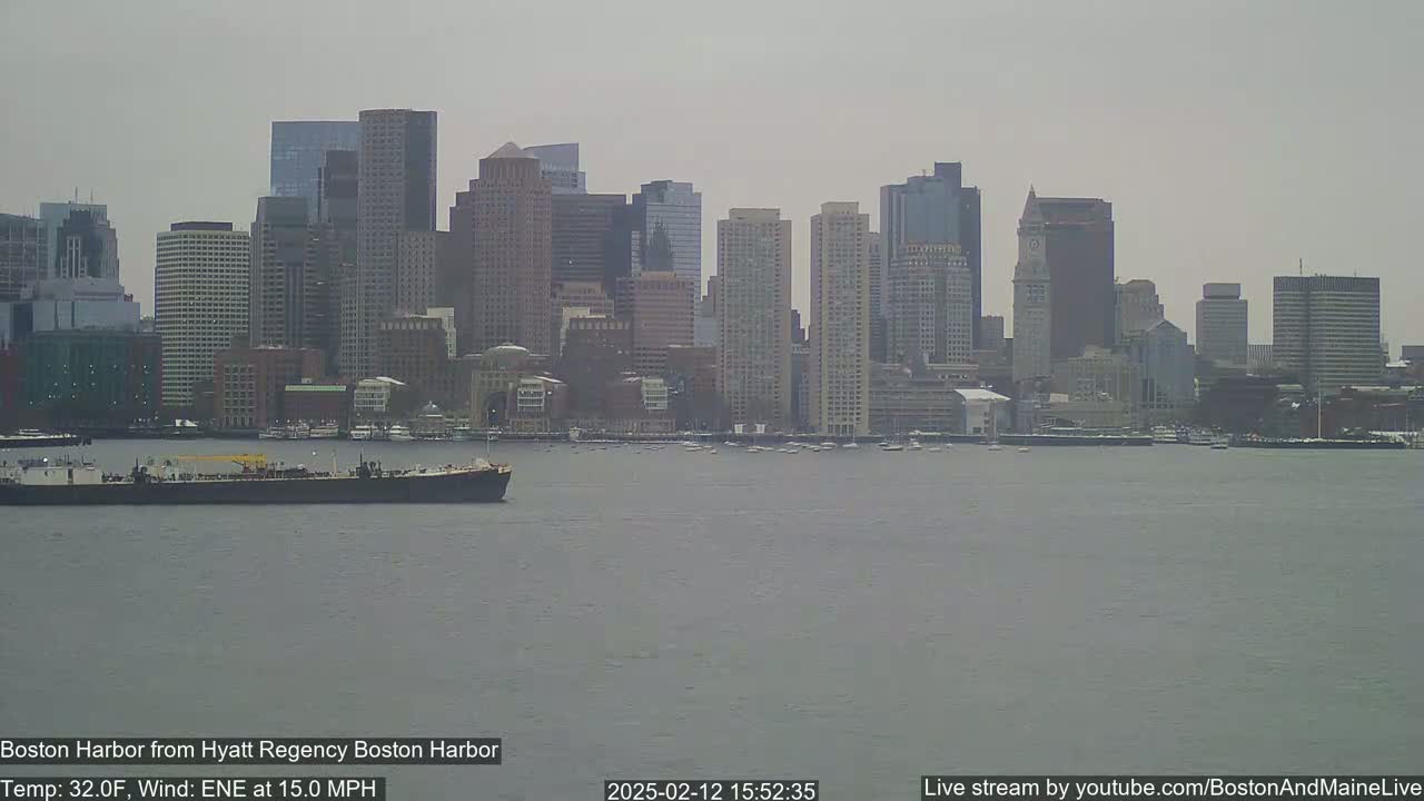 A barge sits in a gray, choppy harbor in front of a city skyline on an overcast day with a 32°F temperature and a 15 mph east-northeast wind.
