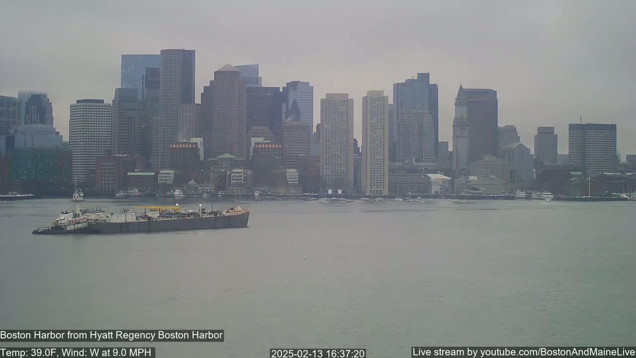A barge is seen on calm water in front of a city skyline under an overcast sky.