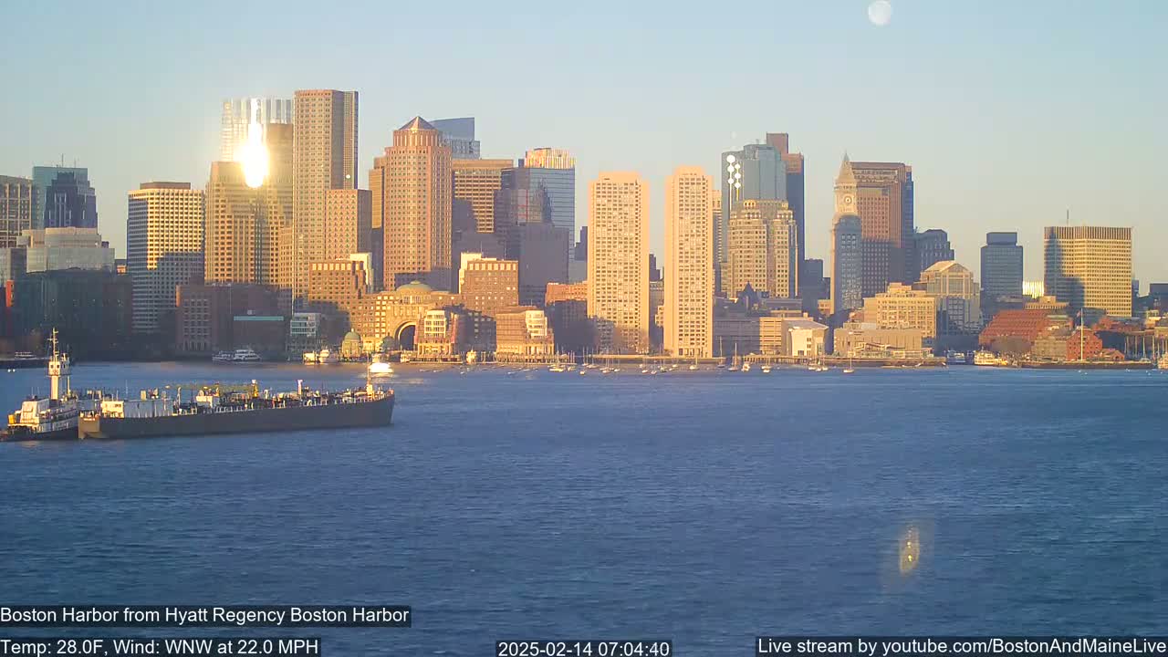A barge is moored in the harbor in front of a city skyline under a clear, sunrise sky.