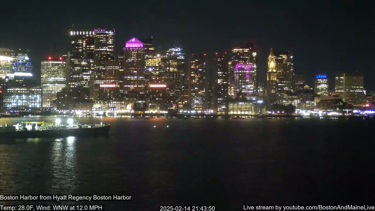 A nighttime view of a brightly lit city skyline across a dark body of water, under a clear sky with a temperature of 28°F and a 12 mph wind.