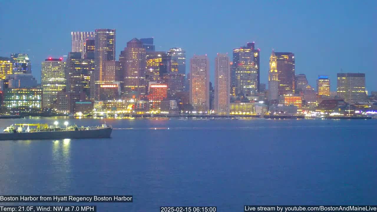 A nighttime view of a city skyline across a dark-blue harbor, with a ship in the foreground and a calm, dark-blue water under a twilight sky.