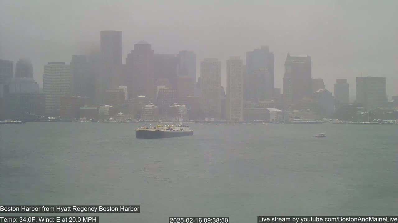 A barge travels across a harbor in front of a hazy city skyline on a cold, windy, and snowy day.