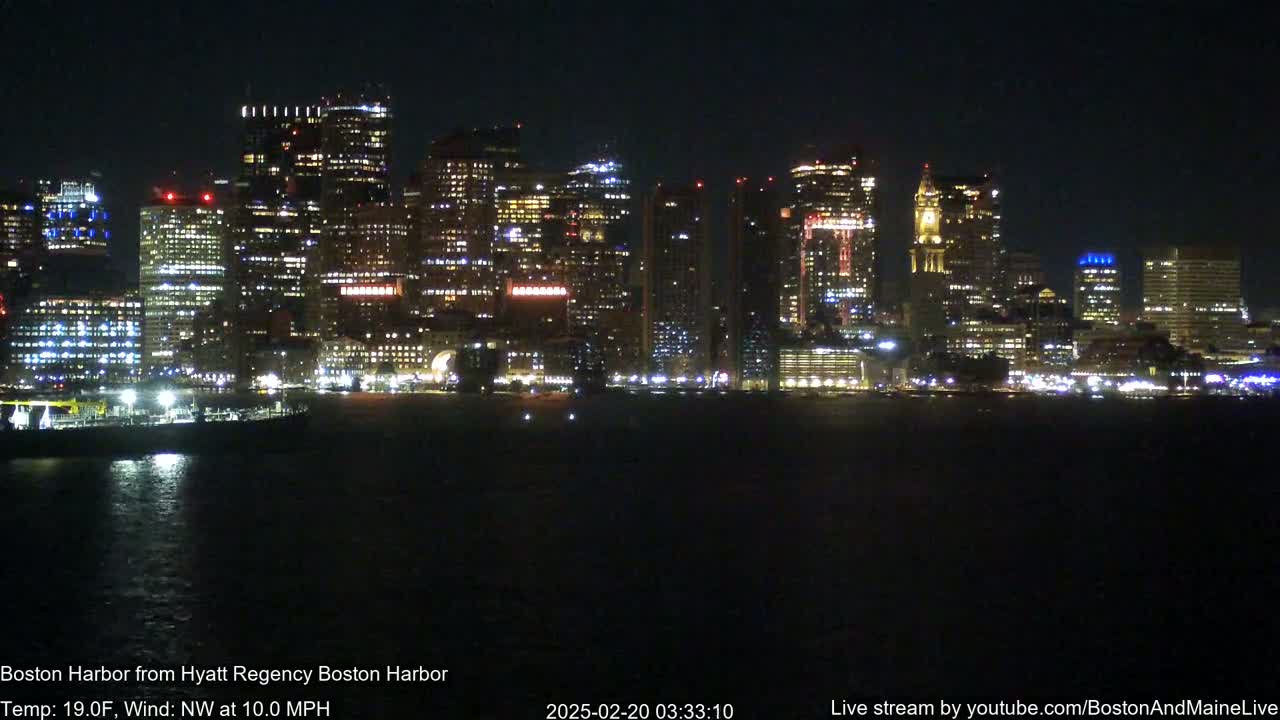 A brightly lit city skyline is seen across a dark body of water at night, with a northwest wind blowing at 10 mph and a temperature of 19°F.