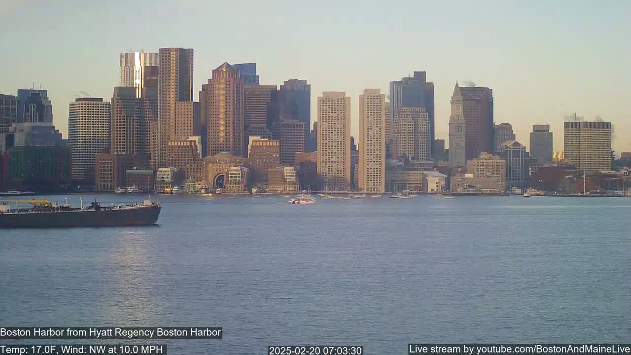 A large cargo ship is in the foreground of a calm body of water with the Boston skyline visible in the background under a clear, pale sky.
