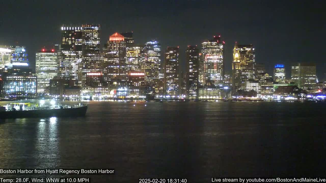A brightly lit city skyline is seen across a dark body of water at night, with a vessel in the foreground and a 10 mph wind.