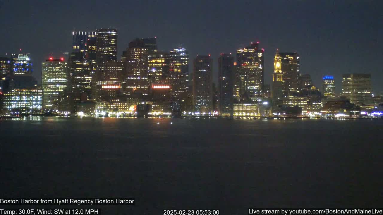 A brightly lit city skyline is seen across a dark body of water at night, under a 30°F temperature with a southwest wind at 12 mph.