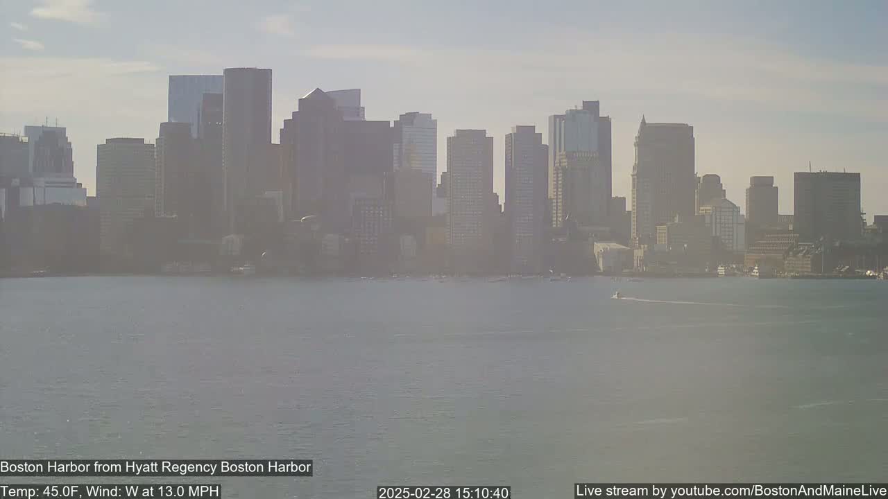 A hazy view of the Boston skyline across a calm harbor on a partly cloudy day.