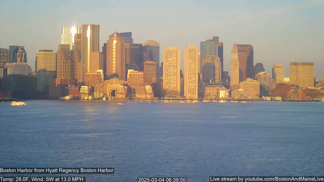A cityscape of numerous skyscrapers and high-rise buildings is seen across a calm body of water under a clear, light-orange sky.