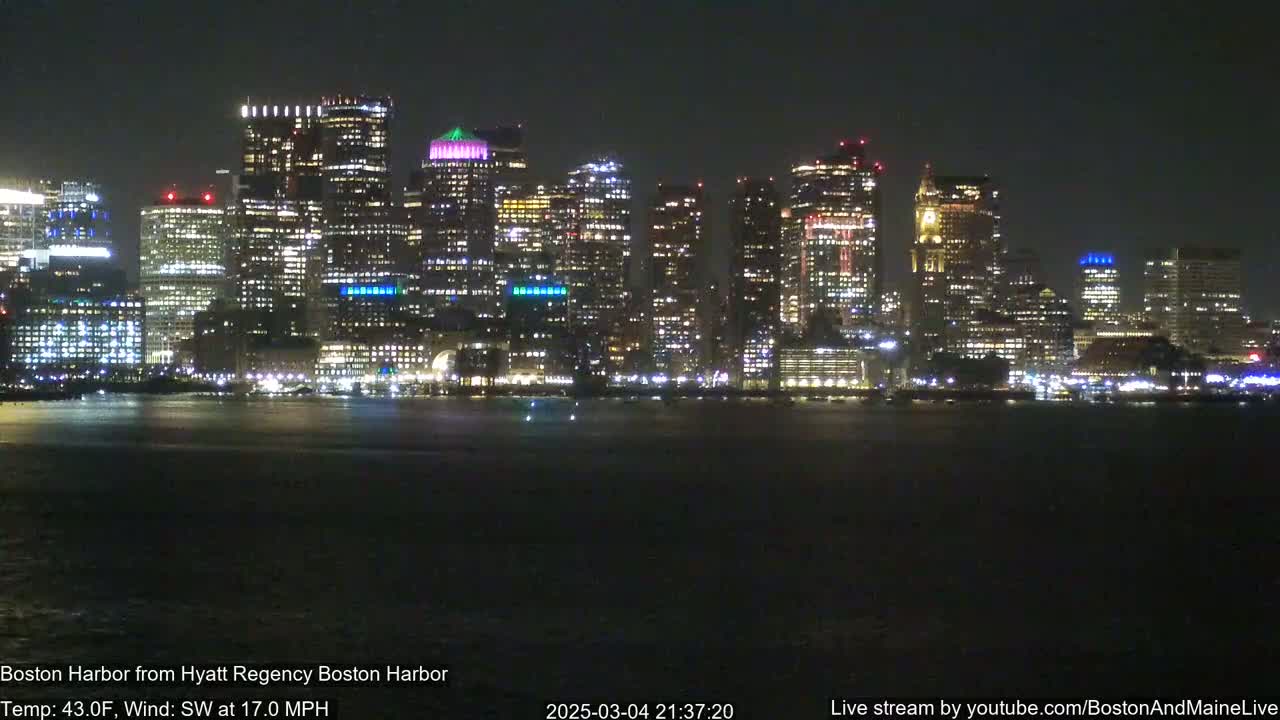 A brightly lit city skyline is seen across a dark body of water at night, under a 43°F temperature with a southwest wind at 17 mph.