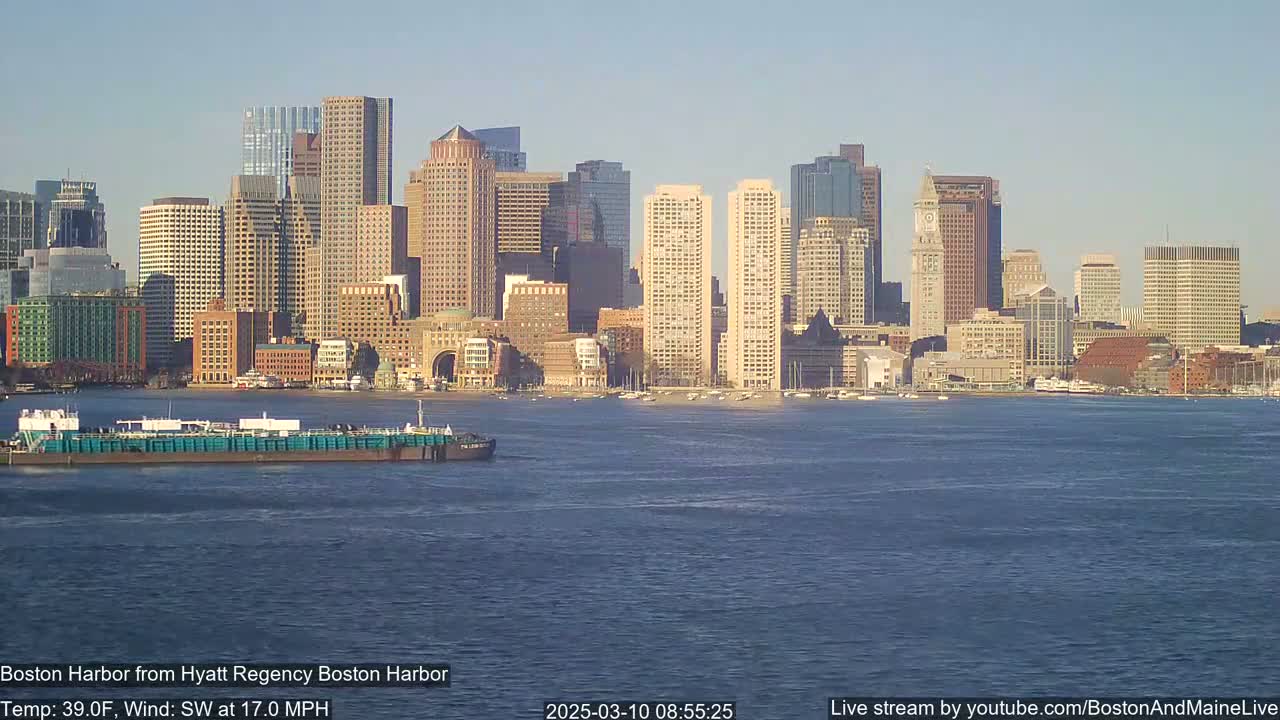 A barge floats on choppy water in front of a city skyline under a clear, sunny sky.