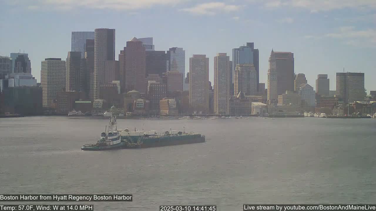 A tugboat pushes a barge across a harbor in front of a city skyline on a partly cloudy day with a west wind at 14 mph and a temperature of 57°F.