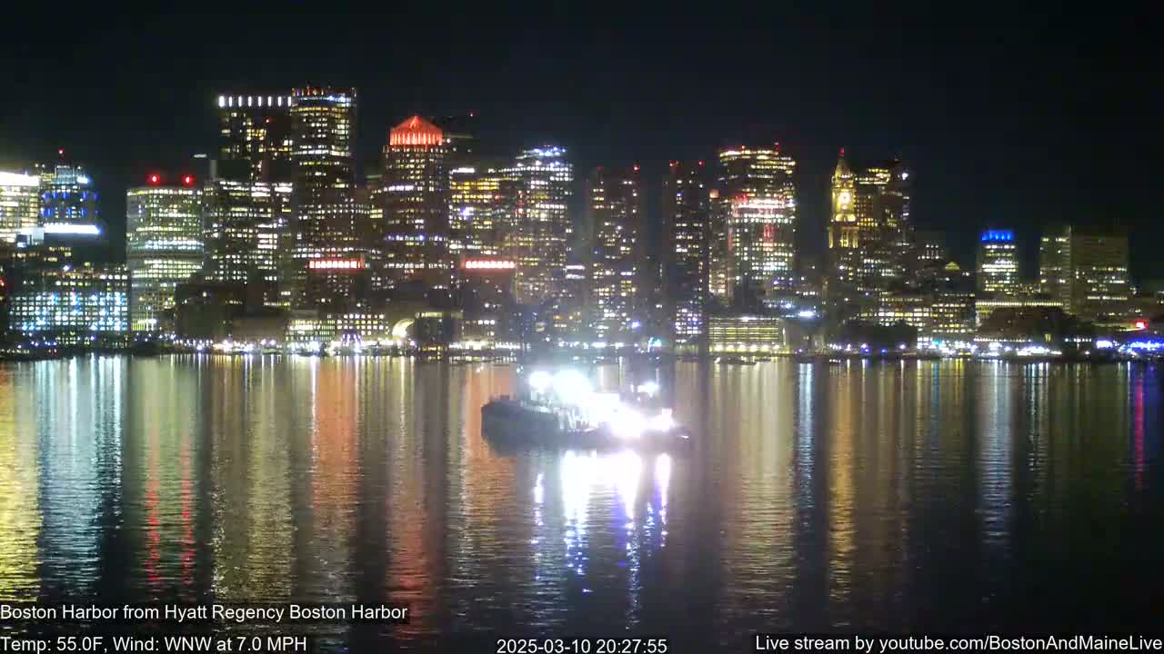 A brightly lit cityscape is reflected in calm nighttime water, with a small boat illuminated in the foreground.